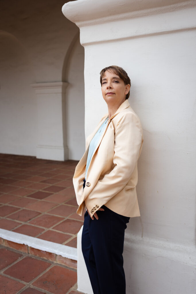 Kathleen is wearing a suit jacket and pants, leaning against a plaster mission style pillar with terracotta floor tile in the background.