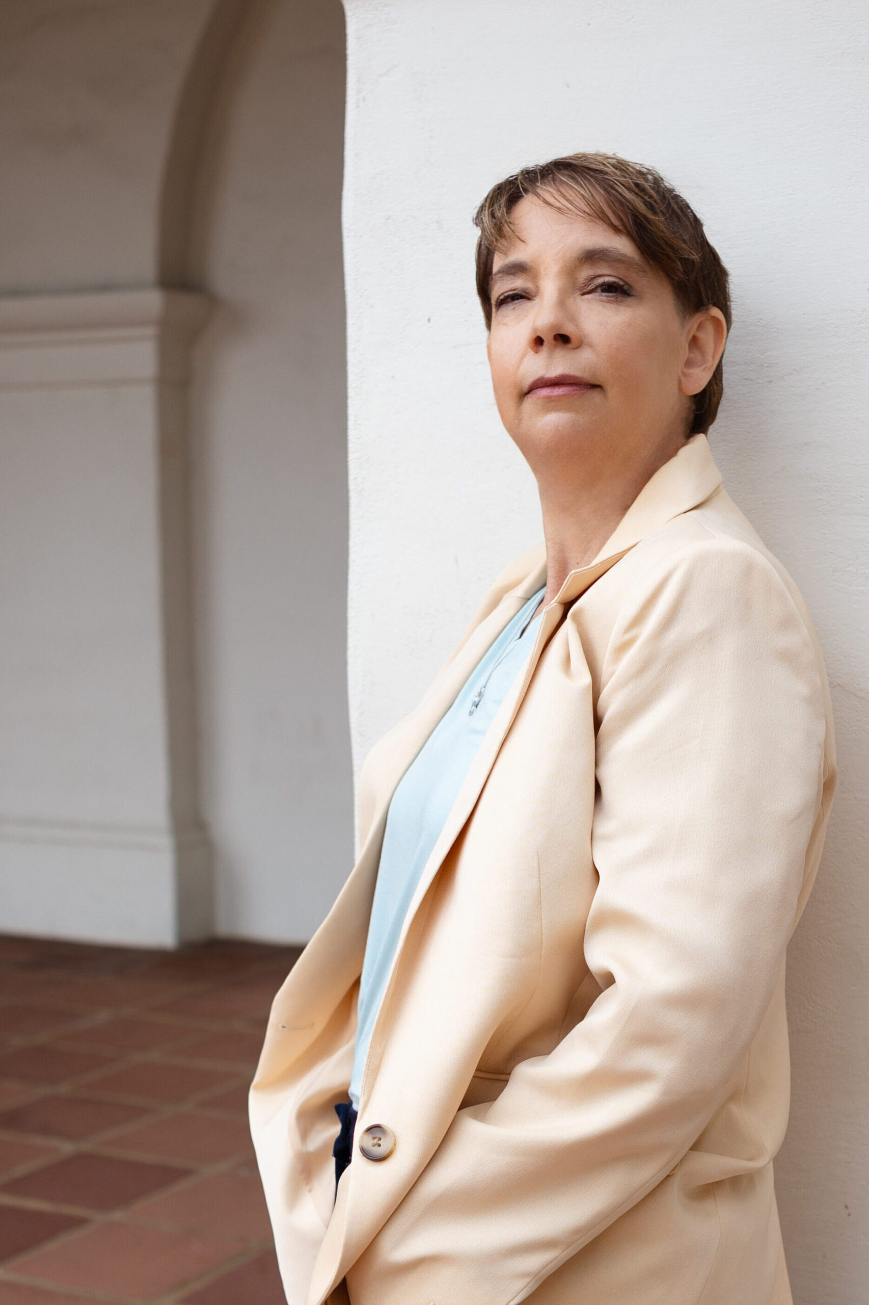 Kathleen is wearing a suit jacket and pants, leaning against a plaster mission style pillar with terracotta floor tile in the background.