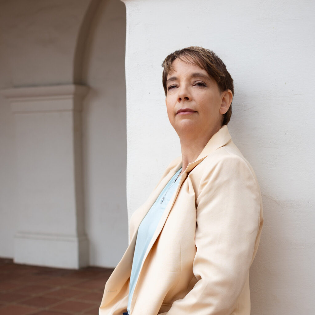 Kathleen is wearing a suit jacket and pants, leaning against a plaster mission style pillar with terracotta floor tile in the background.