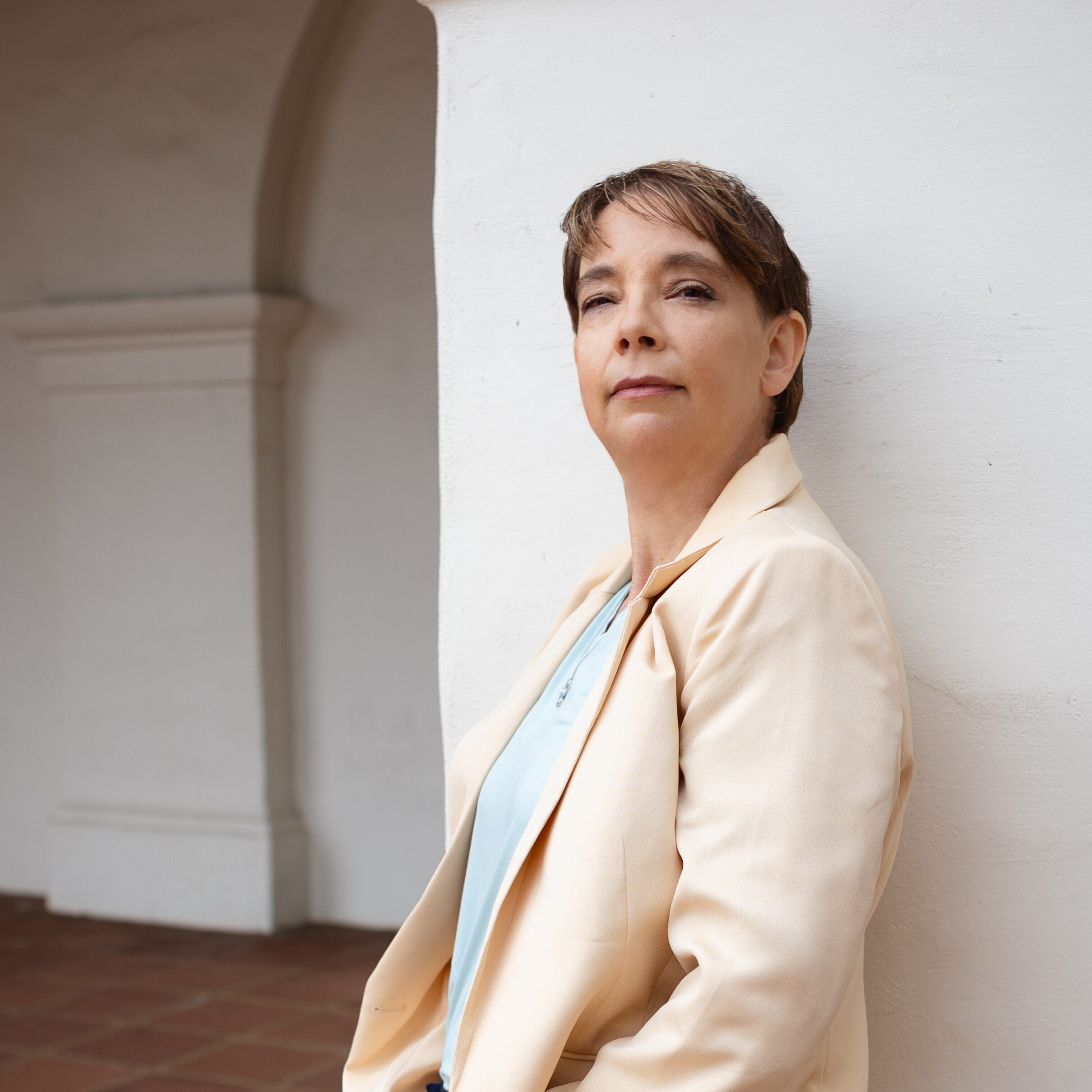 Kathleen is wearing a suit jacket and pants, leaning against a plaster mission style pillar with terracotta floor tile in the background.